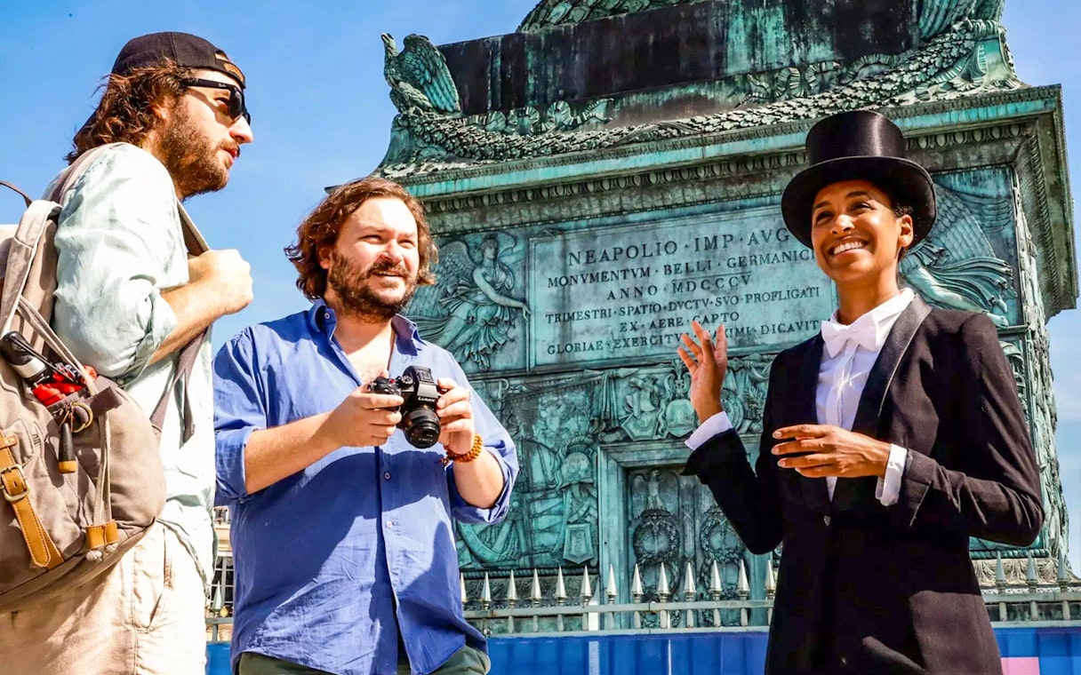 Tour guide in top hat leading Josephine Baker's Paris tour at historical monument.