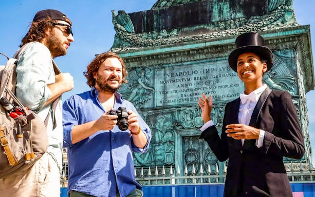 Tour guide in top hat leading Josephine Baker's Paris tour at historical monument.