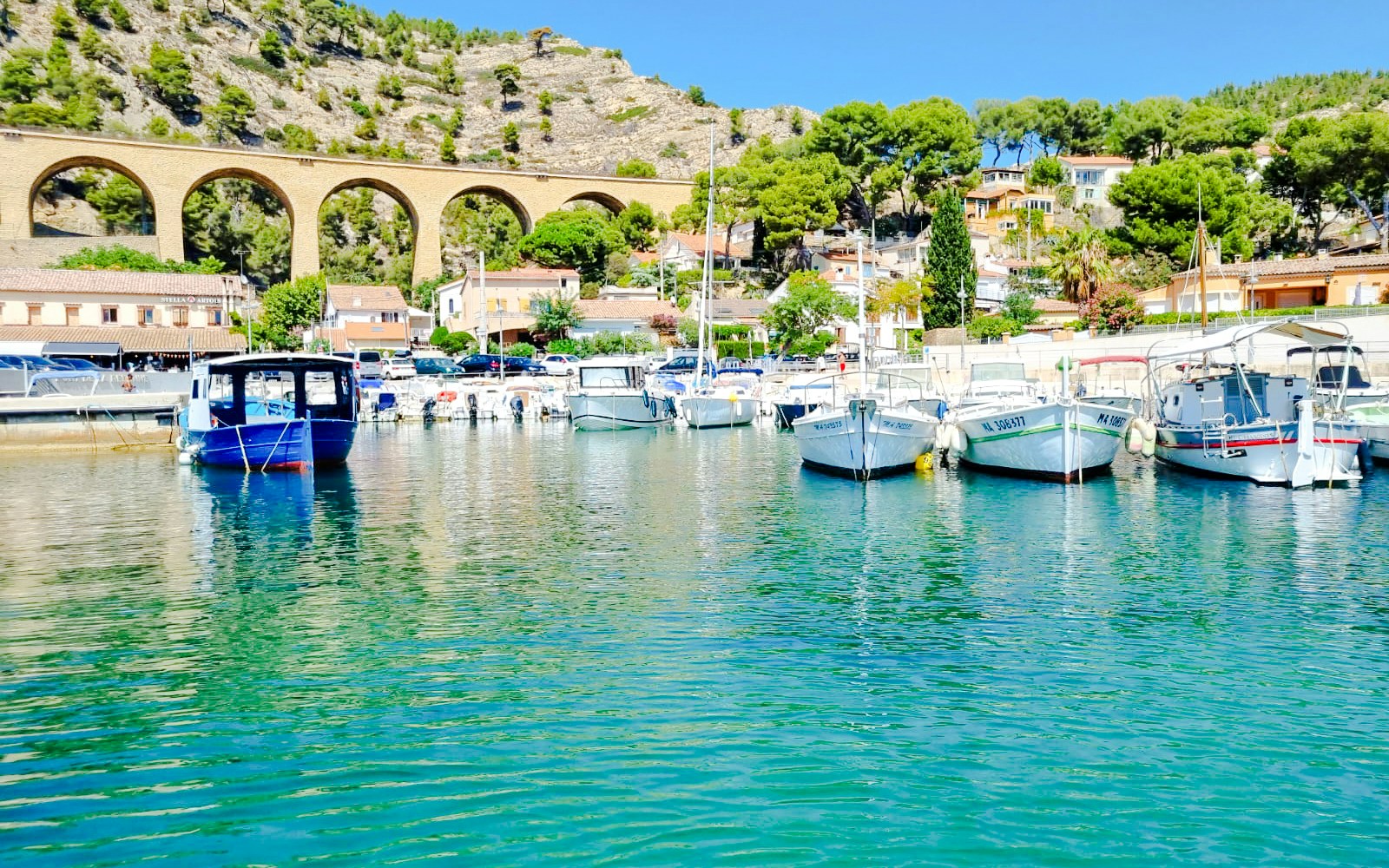 Boats docked in Marseille with a stone viaduct and hillside houses in the background.