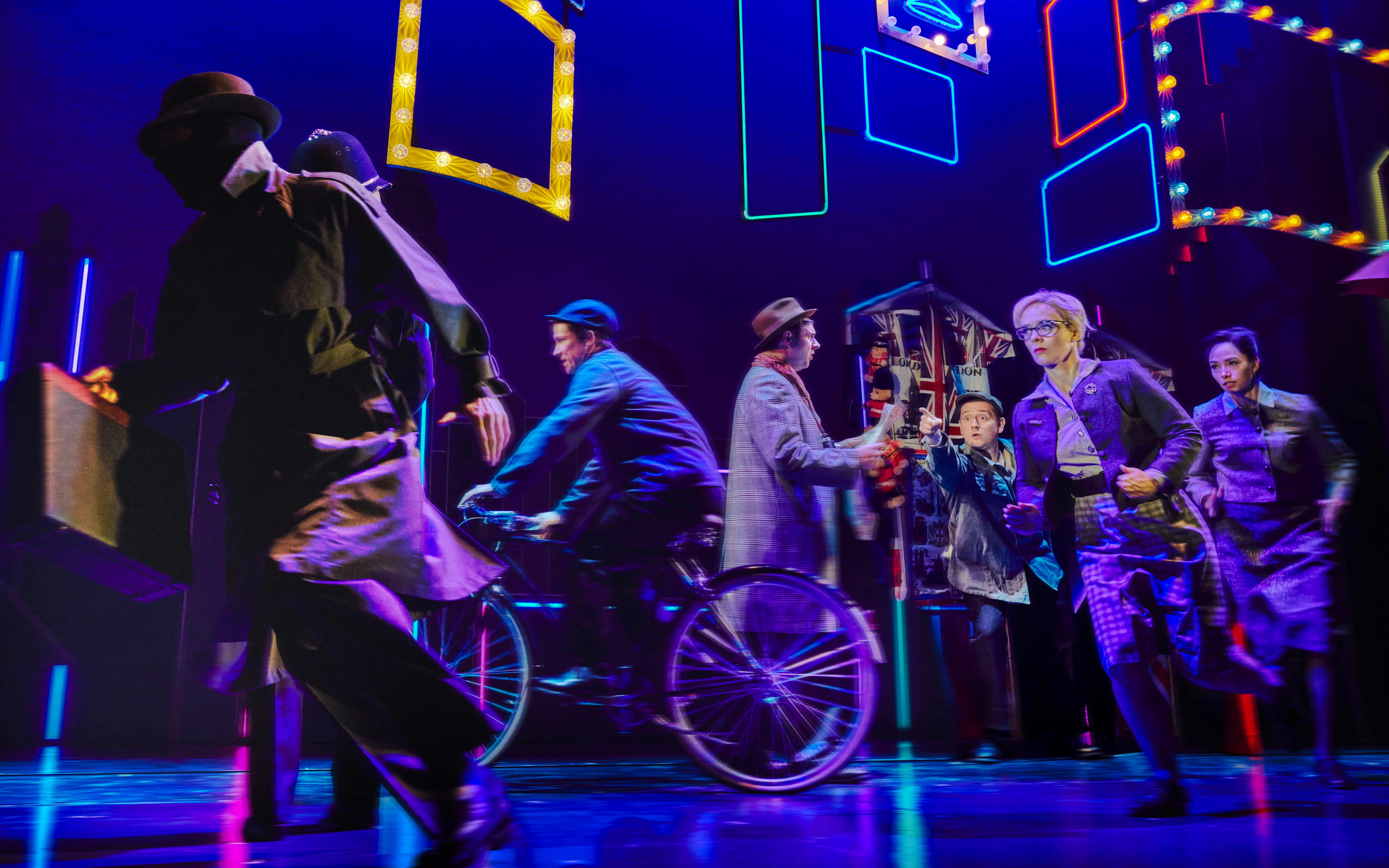 Actors in spy-themed costumes perform energetically on stage with neon lights.