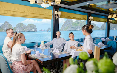Guests enjoy drinks served by a waitress inside a cruise ship dining room overlooking Ha Long Bay.