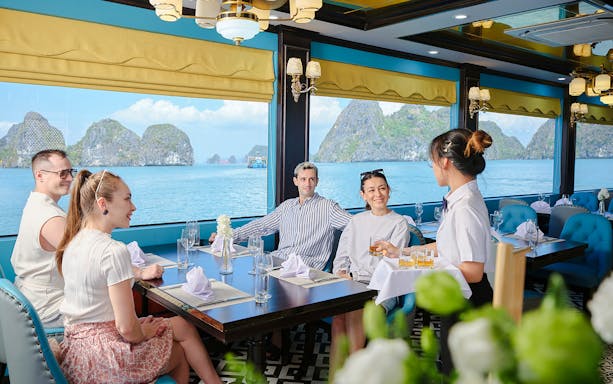 Guests enjoy drinks served by a waitress inside a cruise ship dining room overlooking Ha Long Bay.
