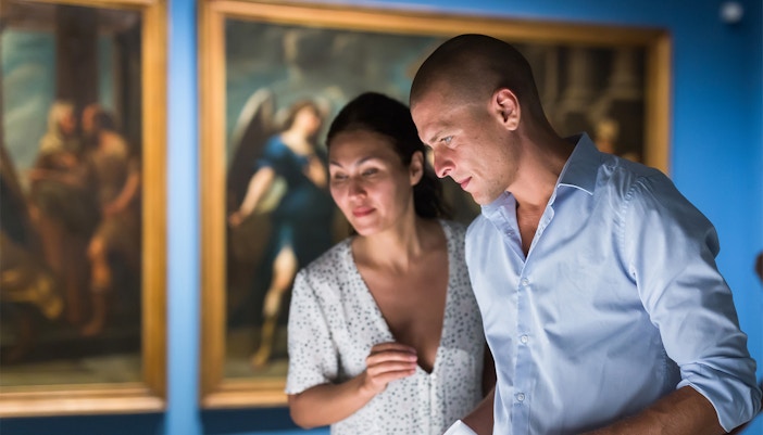 The image shows a man and a woman looking at paintings at an art gallery.