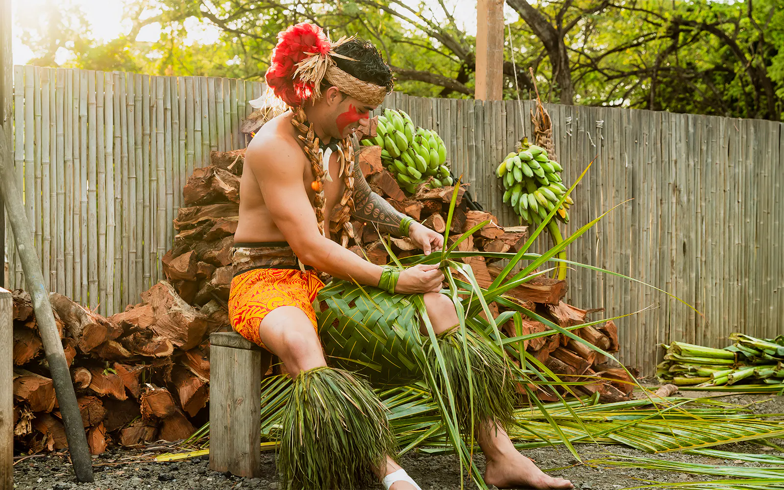 Man weaving palm fronds at Mauka Warriors Luau, surrounded by bananas and wood.