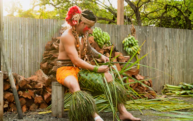 Man weaving palm fronds at Mauka Warriors Luau, surrounded by bananas and wood.