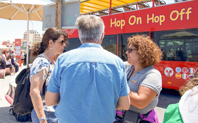 Heraklion tourists discussing with guide near Hop-On Hop-Off bus.