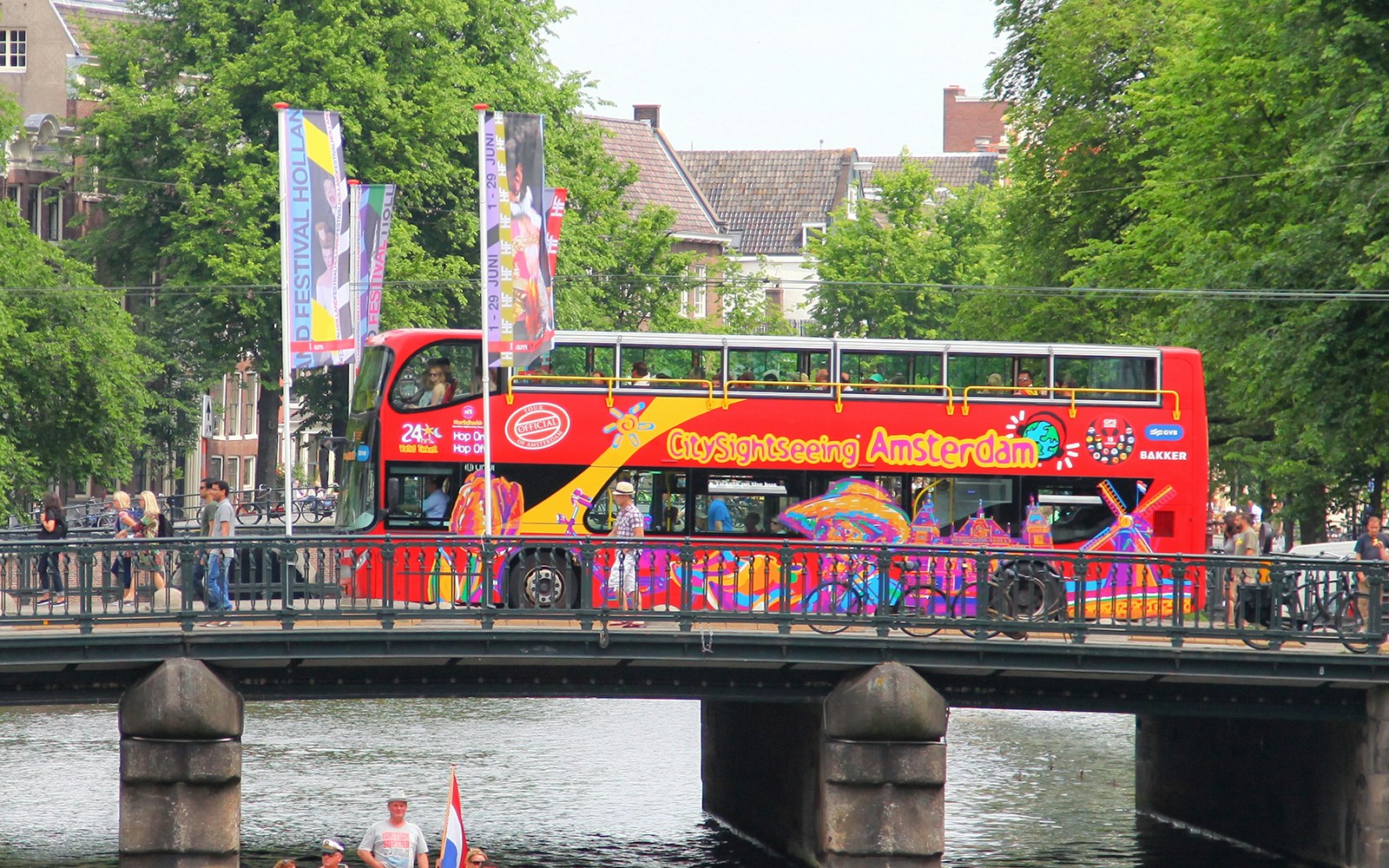 Amsterdam sightseeing bus crossing canal bridge with historic buildings in background.