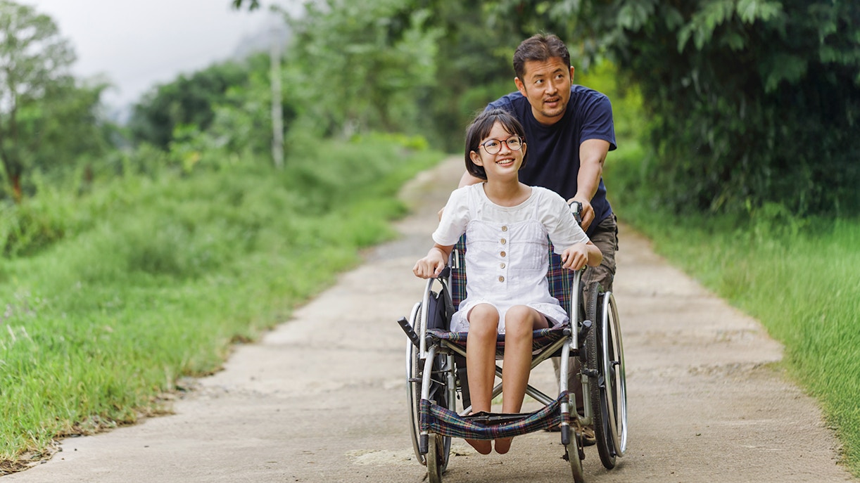 Man pushing a girl in a wheelchair through a scenic park in New York City.