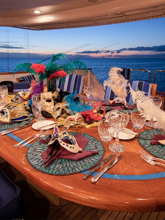 Dinner cruise table set with Venetian masks and crystal glasses at sunset.