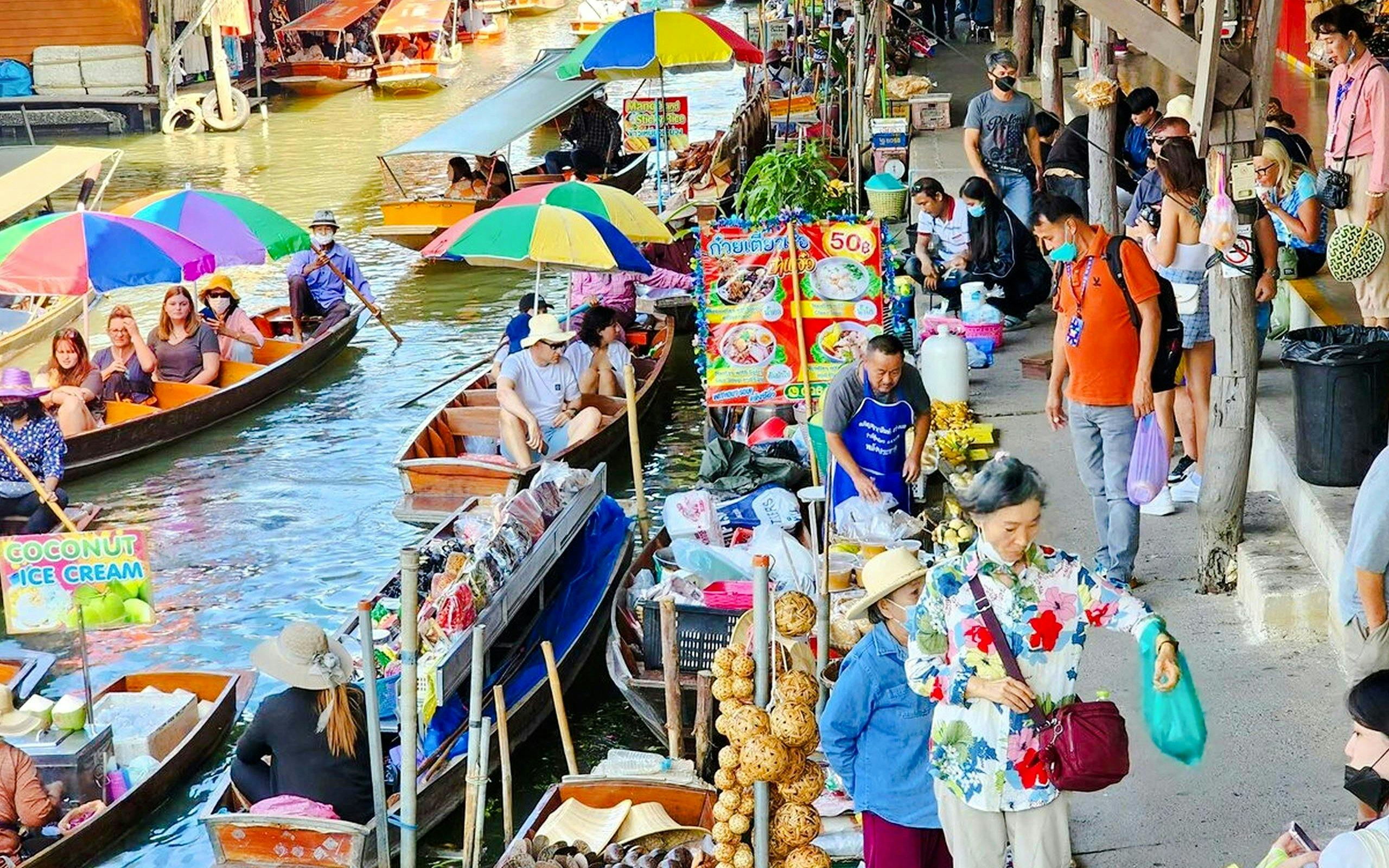 Tourists navigating boats at Damnoen Saduak Floating Market, Thailand, with colorful stalls.