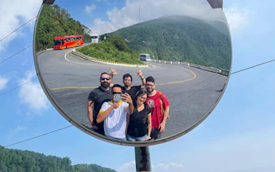 Tourists taking a selfie in a convex mirror on Hai Van Pass, Vietnam, with scenic mountain road.