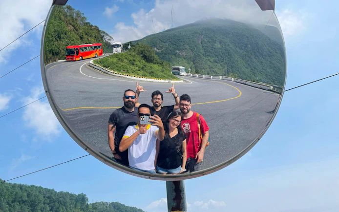 Tourists taking a selfie in a convex mirror on Hai Van Pass, Vietnam, with scenic mountain road.