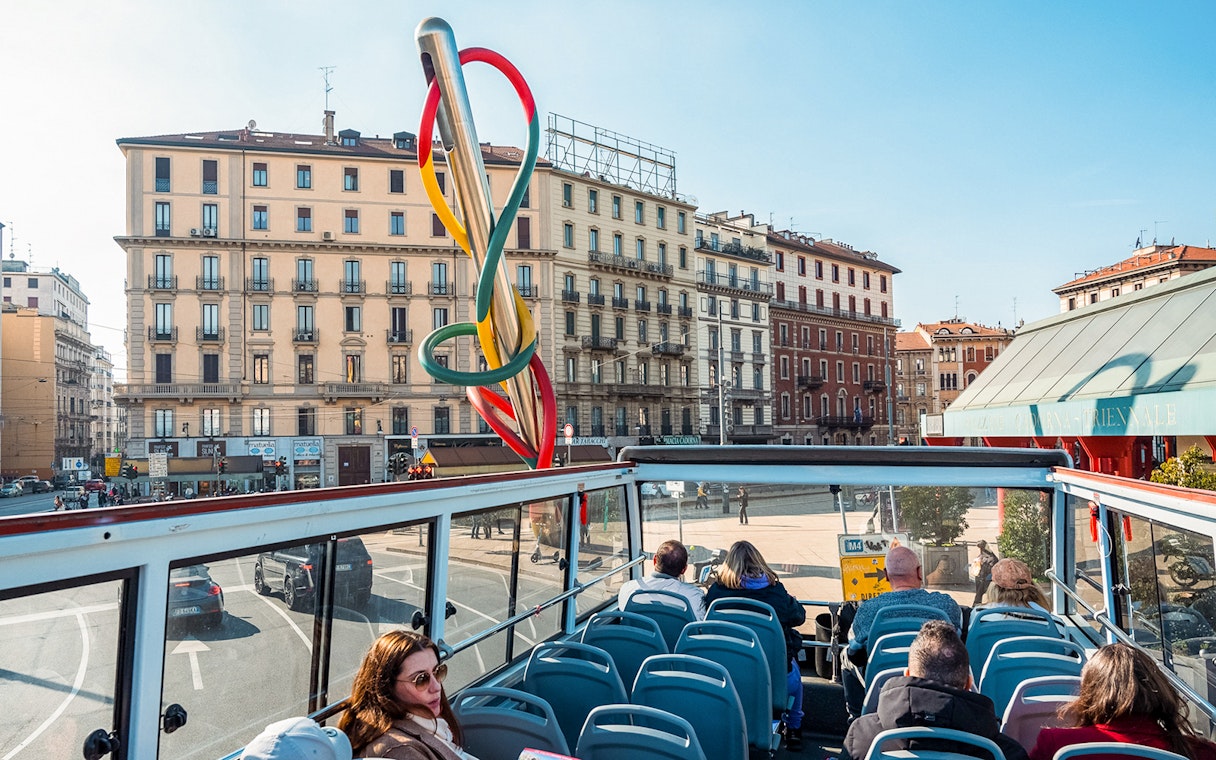 Open-top bus tour in Milan passing by colorful Needle sculpture and historic buildings.