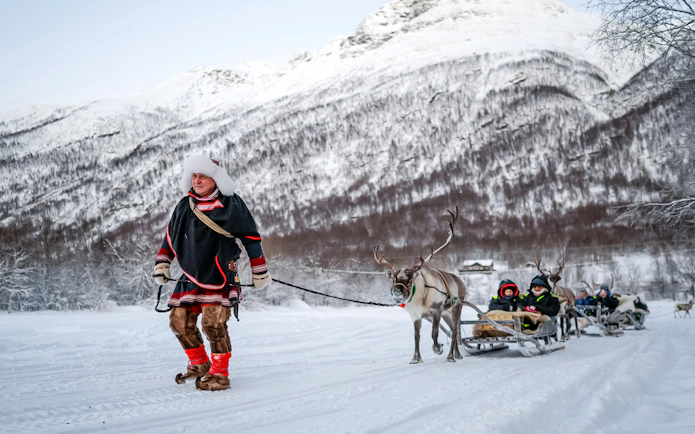 Sámi guide leading reindeer pulling sled with guests through snowy landscape.