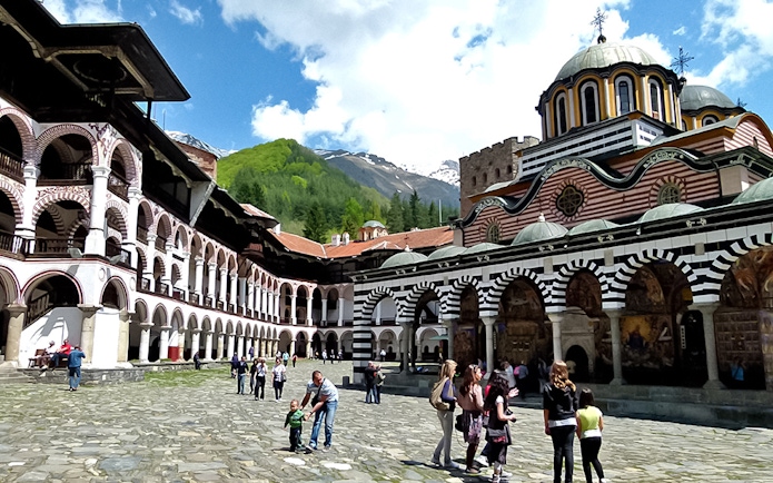 Rila Monastery courtyard with visitors, surrounded by arched buildings and mountain backdrop.