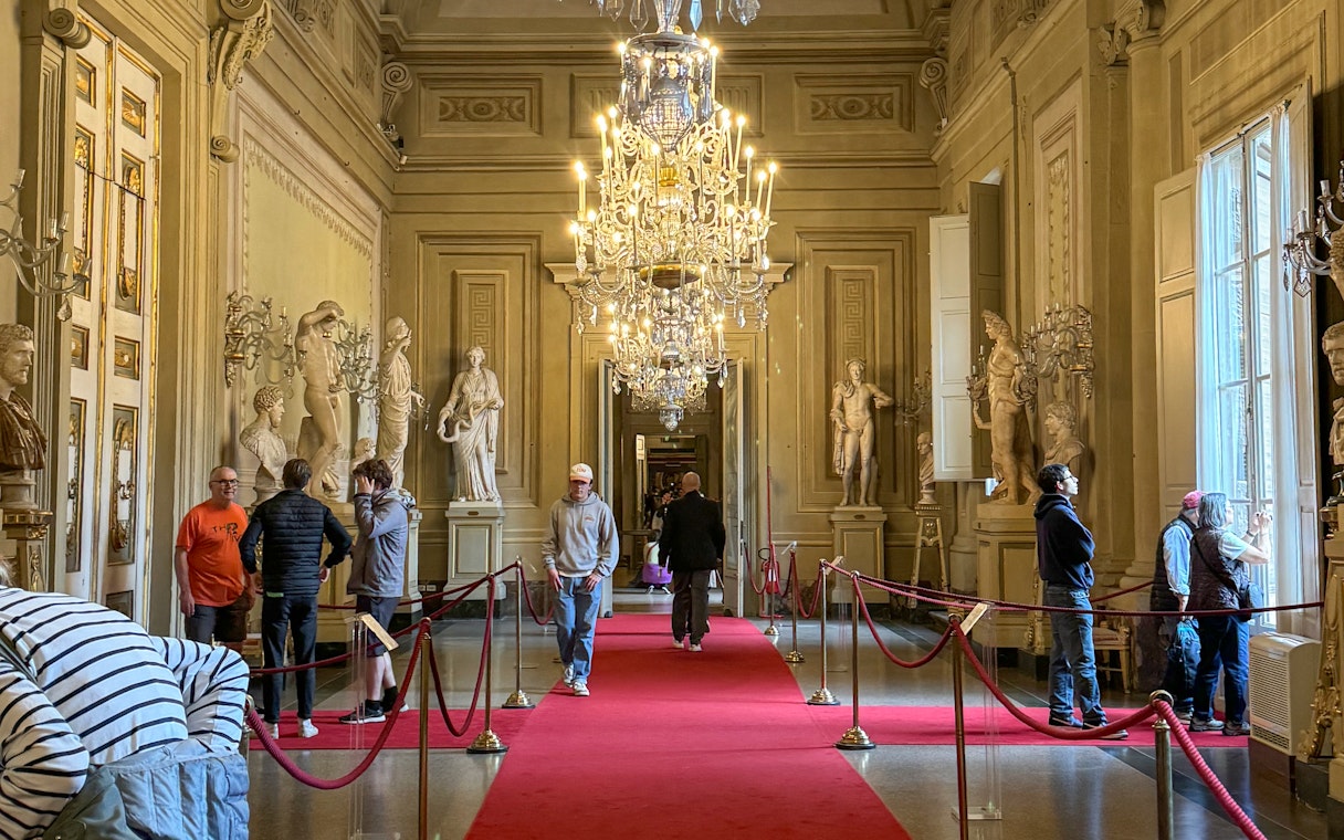 Visitors exploring statues and chandeliers in the Palatine Gallery, Pitti Palace, Florence.