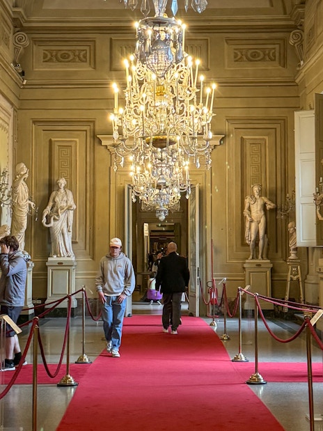 Visitors exploring statues and chandeliers in the Palatine Gallery, Pitti Palace, Florence.