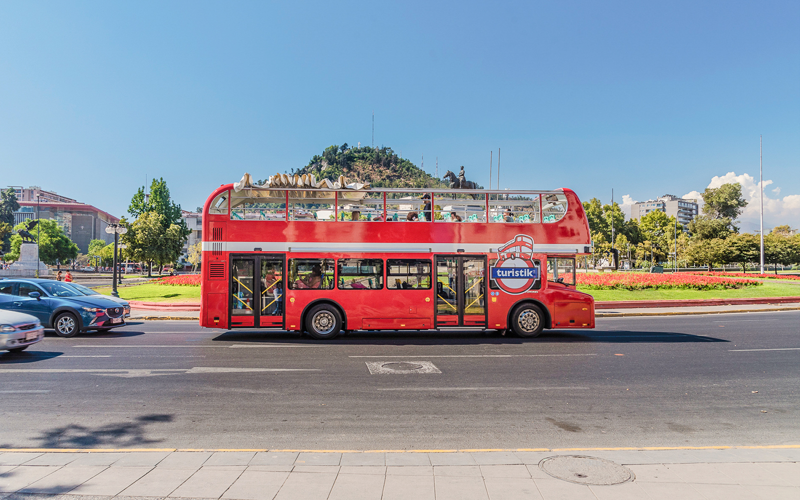 Red double-decker bus on Santiago Hop-on Hop-off tour with cityscape and hill in background.