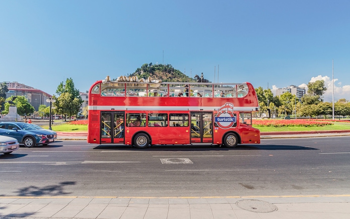 Red double-decker bus on Santiago Hop-on Hop-off tour with cityscape and hill in background.