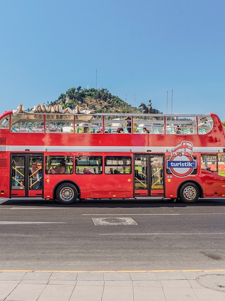 Red double-decker bus on Santiago Hop-on Hop-off tour with cityscape and hill in background.