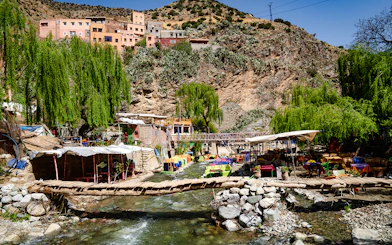 Wooden bridge over a stream with cafes and colorful tables in Ourika Valley, Morocco.