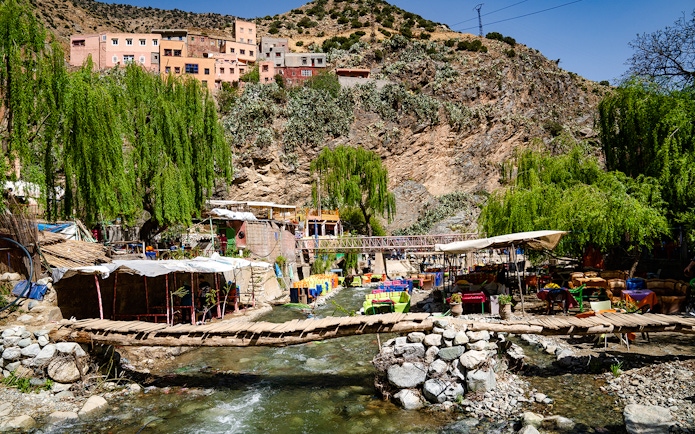 Wooden bridge over a stream with cafes and colorful tables in Ourika Valley, Morocco.