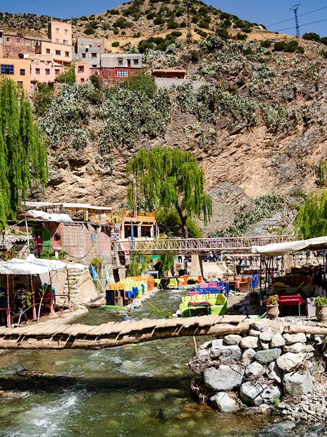Wooden bridge over a stream with cafes and colorful tables in Ourika Valley, Morocco.