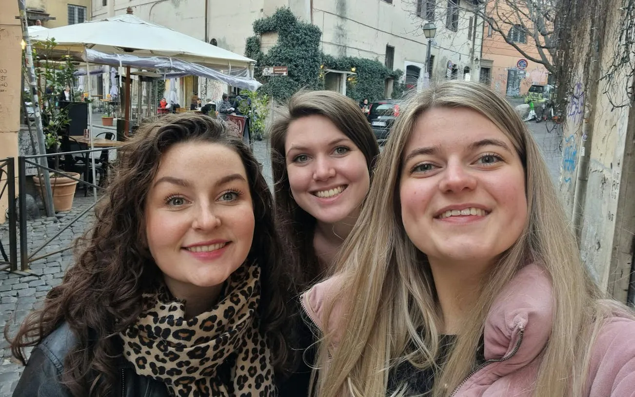 Three people smiling in a cobblestone street in Trastevere, Rome, with outdoor café in the background.