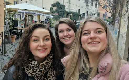 Three people smiling in a cobblestone street in Trastevere, Rome, with outdoor café in the background.
