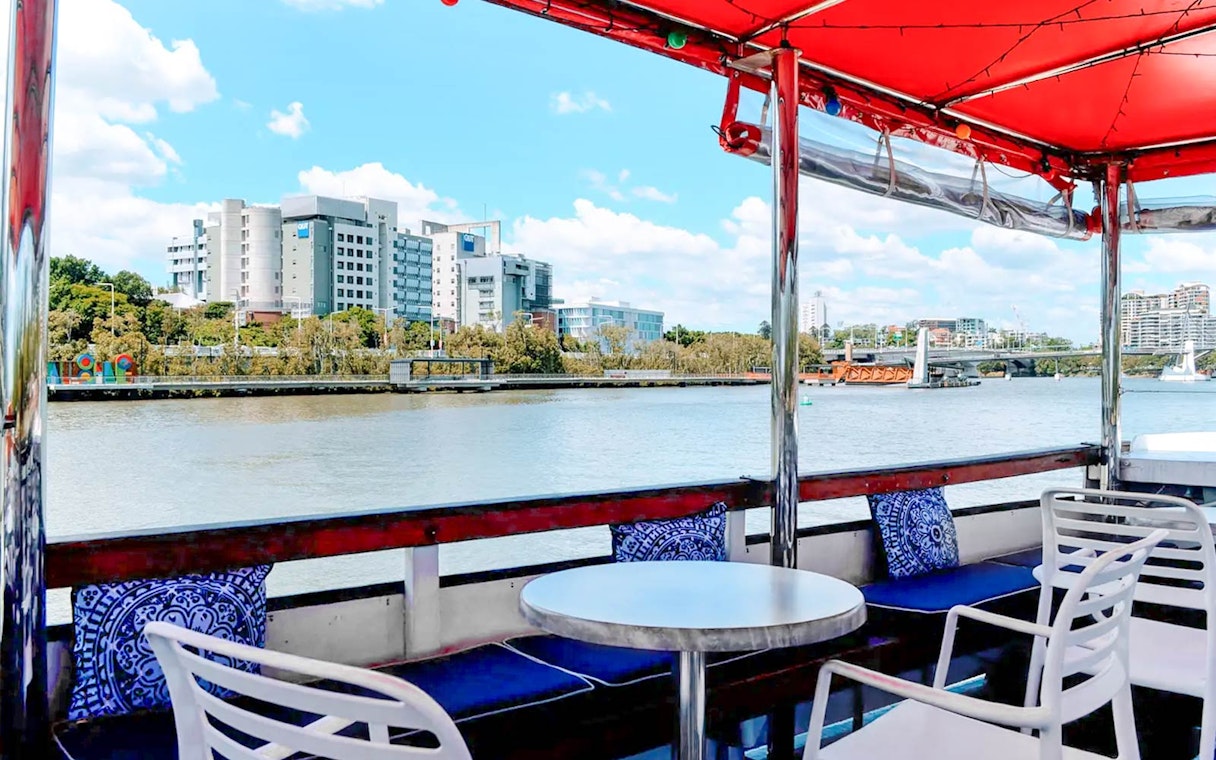 Covered deck view of Brisbane River cruise with city buildings and bridge in the background.