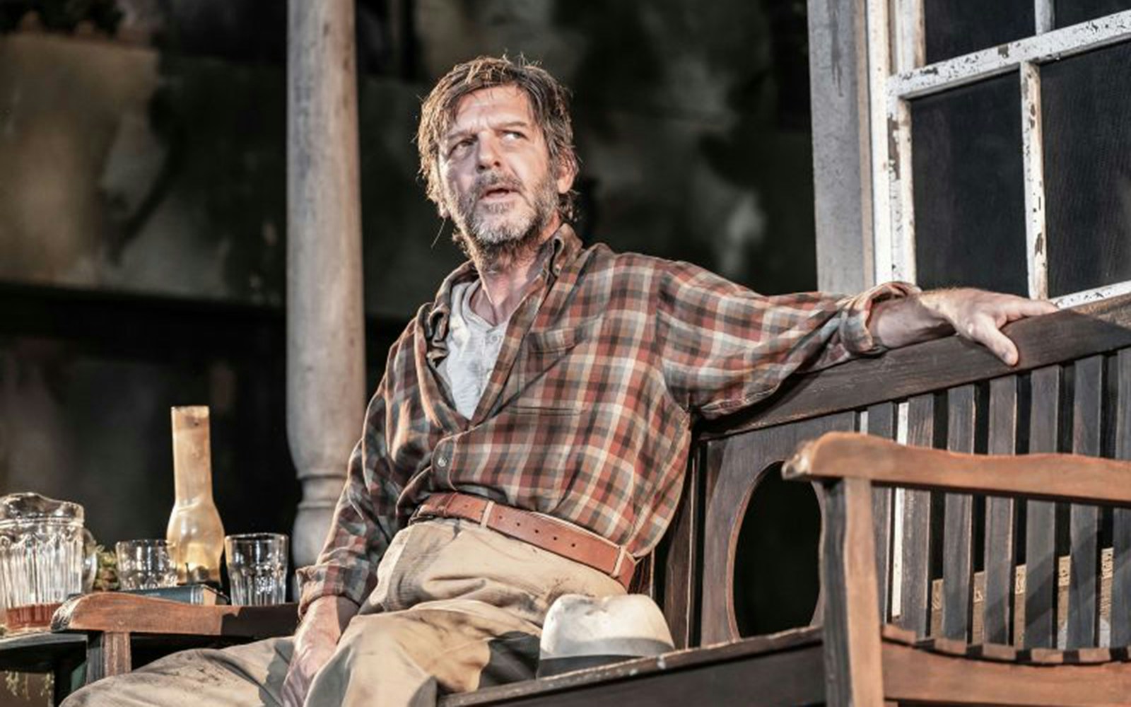 Man in plaid shirt sitting on porch bench, scene from "To Kill A Mockingbird" play.