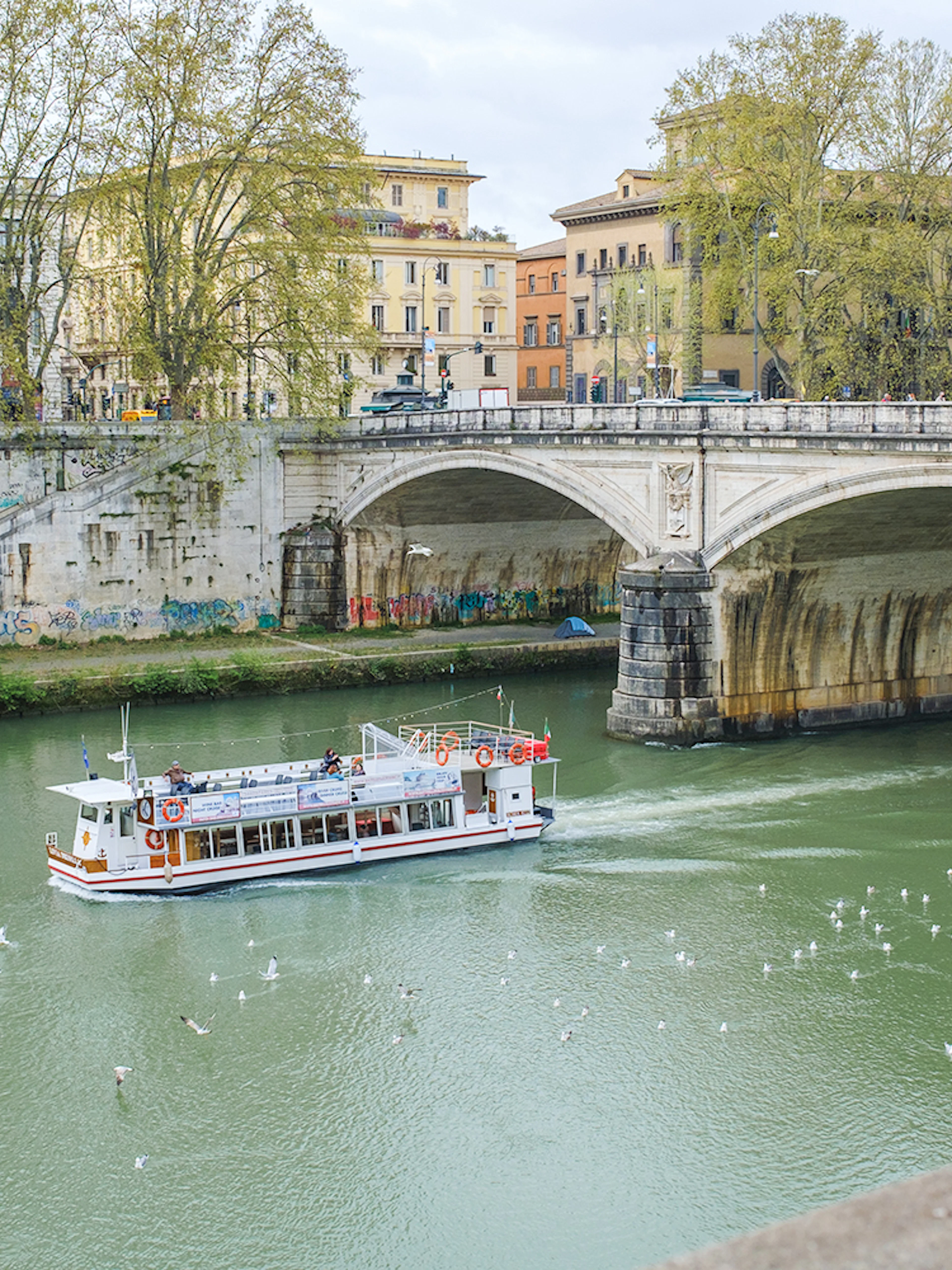 Hop-on hop-off river cruise boat on the Tiber River near a historic bridge in Rome.