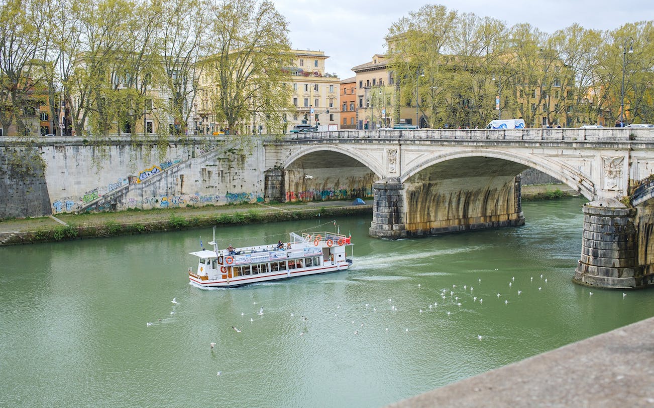 Hop-on hop-off river cruise boat on the Tiber River near a historic bridge in Rome.