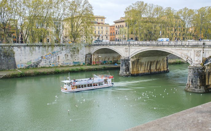 Hop-on hop-off river cruise boat on the Tiber River near a historic bridge in Rome.