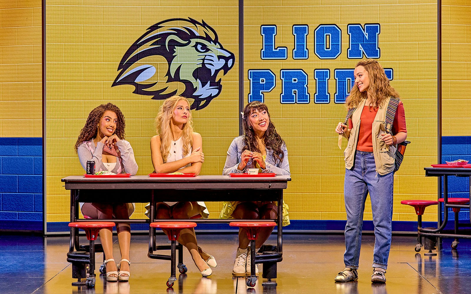Four actors perform a cafeteria scene in Mean Girls The Musical with a "Lion Pride" backdrop.