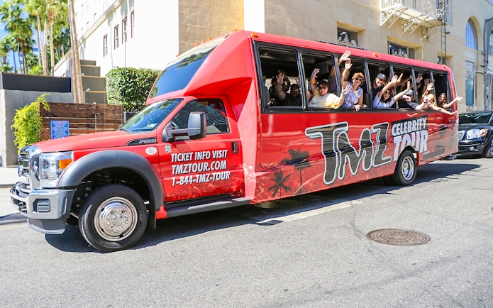 TMZ tour bus with passengers waving on Hollywood street.