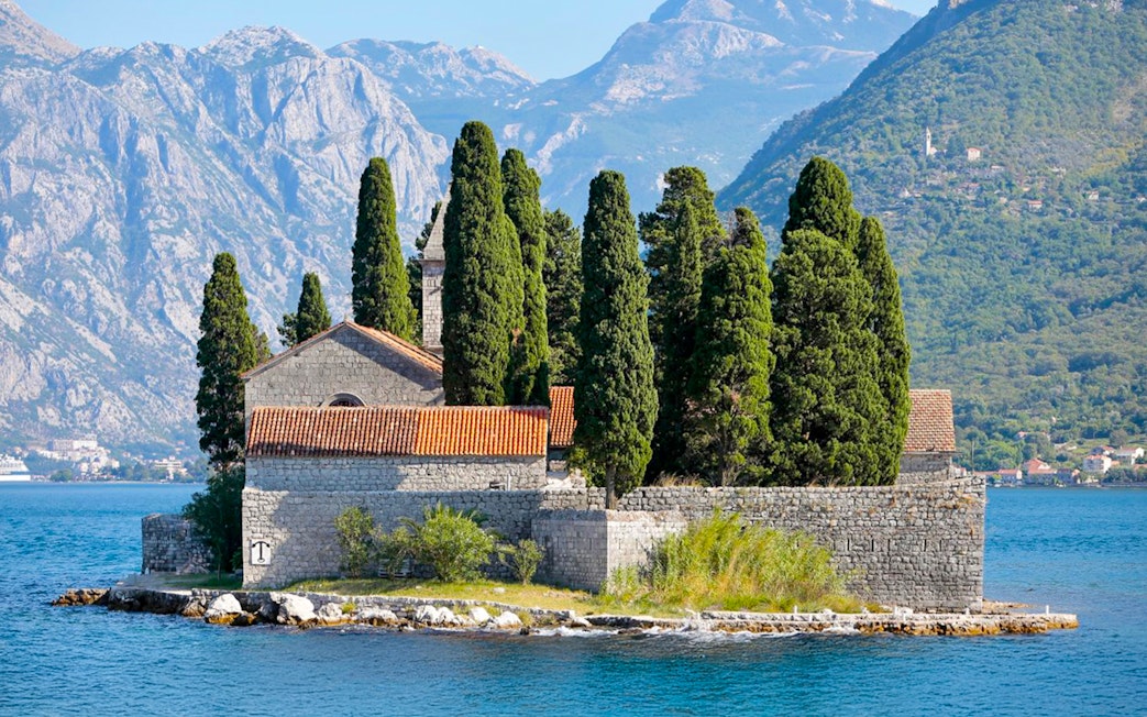 St. George Island in Perast with stone church and cypress trees against mountain backdrop.