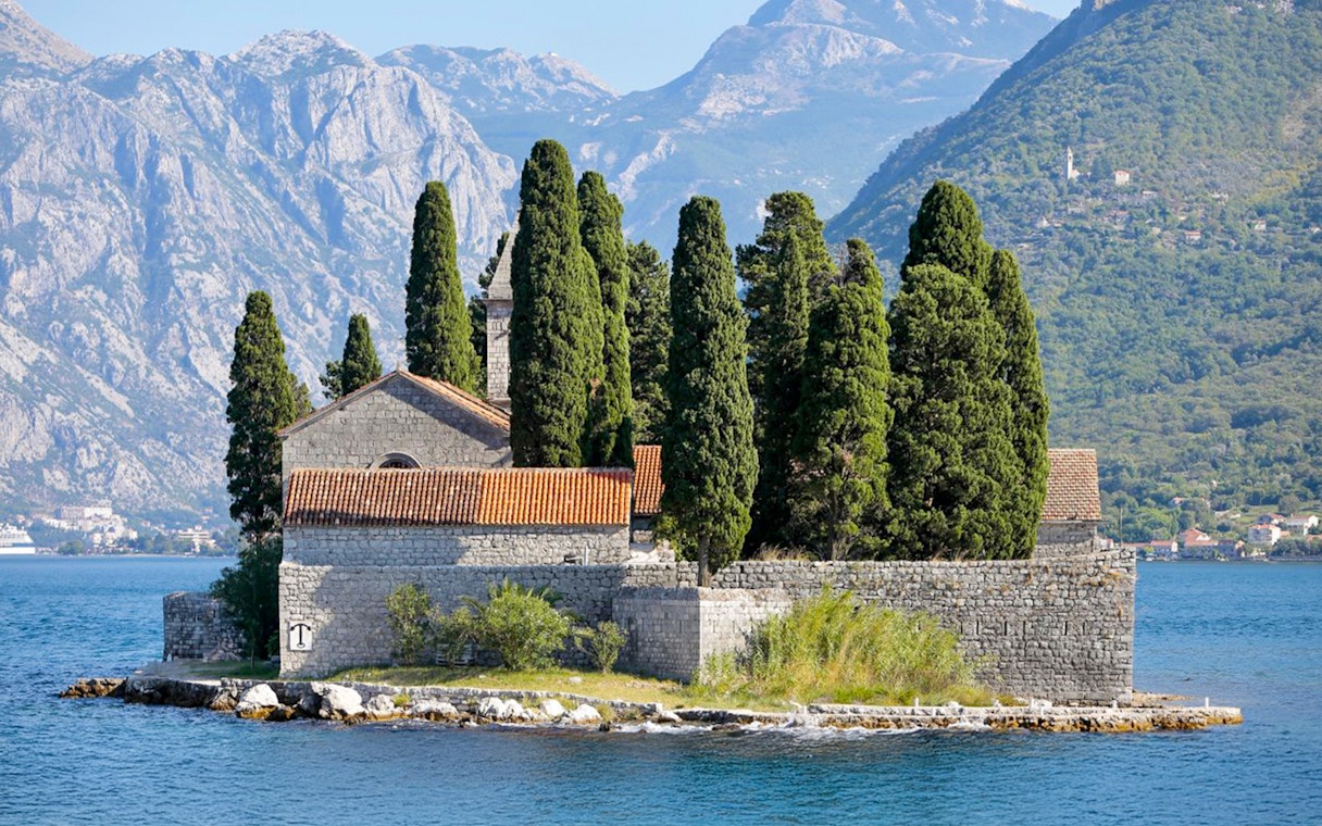 St. George Island in Perast with stone church and cypress trees against mountain backdrop.