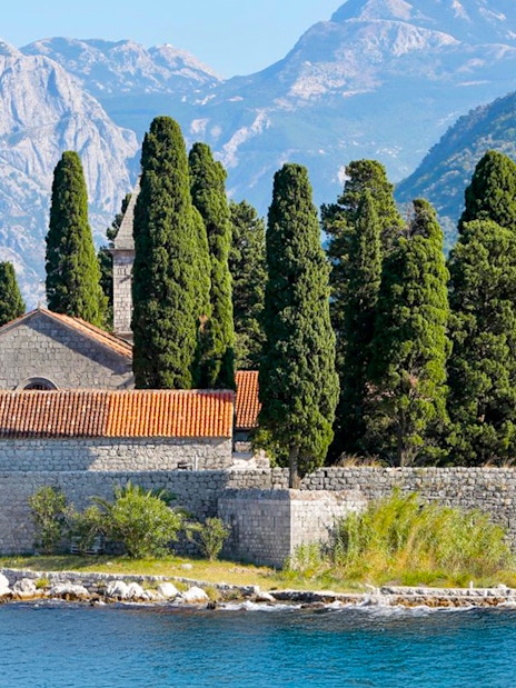 St. George Island in Perast with stone church and cypress trees against mountain backdrop.