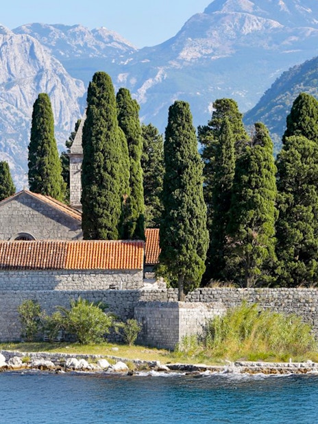 St. George Island in Perast with stone church and cypress trees against mountain backdrop.