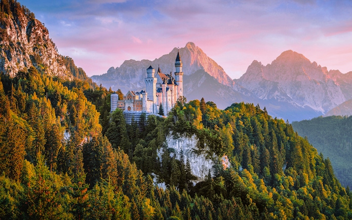 Neuschwanstein Castle aerial view at sunset, surrounded by forested mountains.