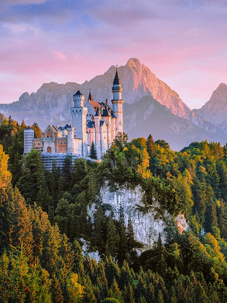 Neuschwanstein Castle aerial view at sunset, surrounded by forested mountains.