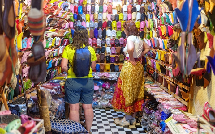 Shoppers browsing colorful slippers in Marrakech souk near Ben Youssef Medersa.