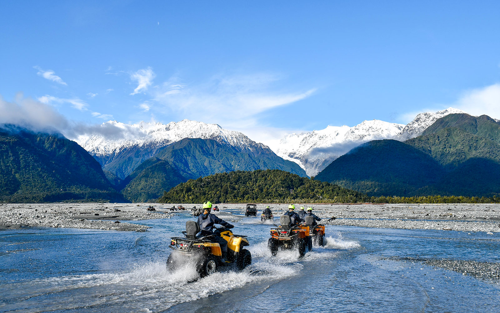 Quad biking through river with snow-capped mountains at Franz Josef, New Zealand.