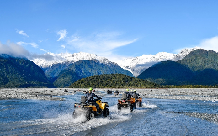 Quad biking through river with snow-capped mountains at Franz Josef, New Zealand.