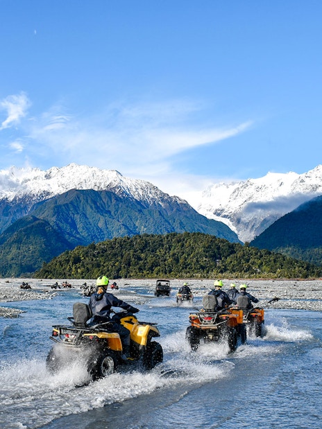 Quad biking through river with snow-capped mountains at Franz Josef, New Zealand.