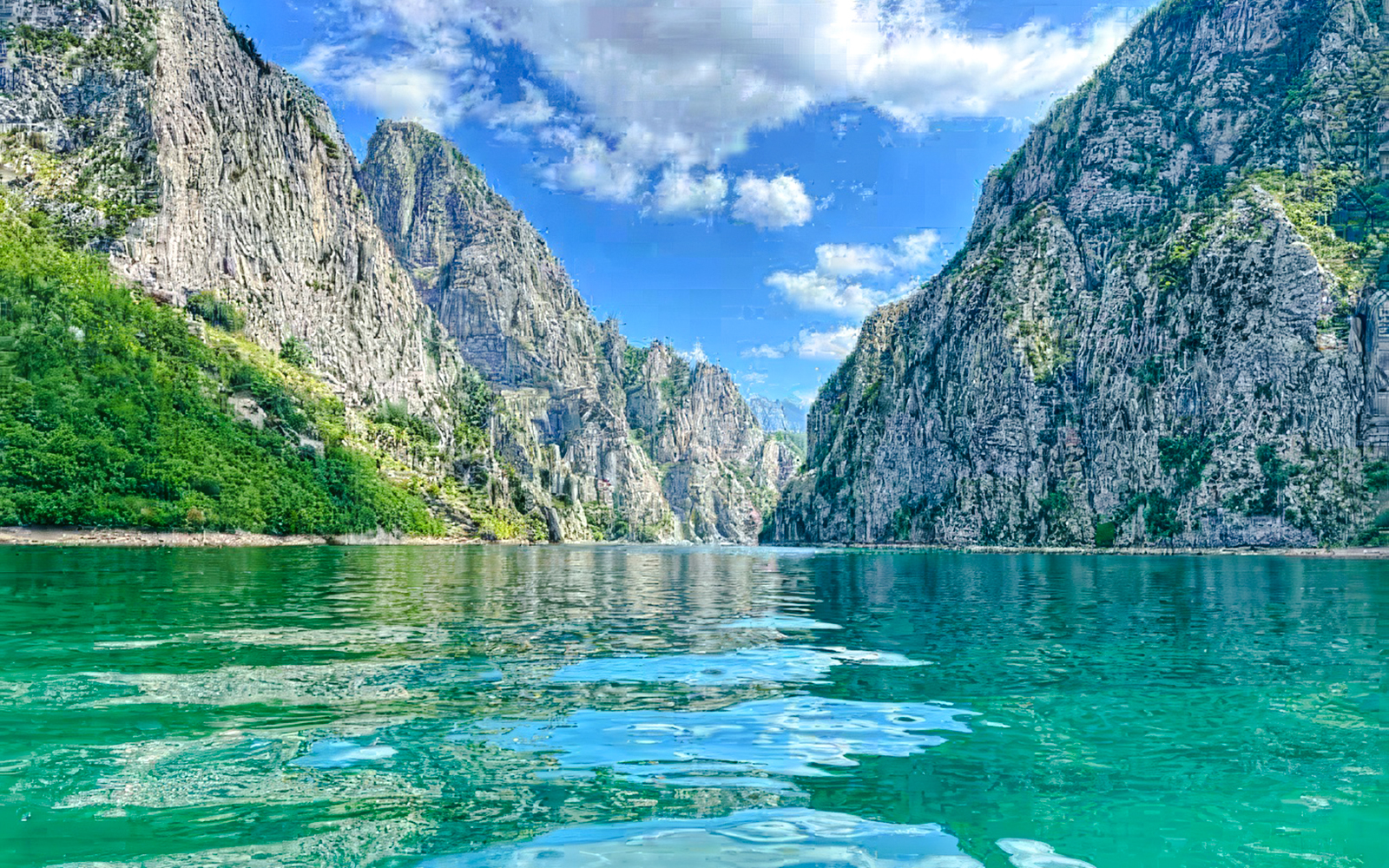 Komani Lake surrounded by steep rocky cliffs under a blue sky.