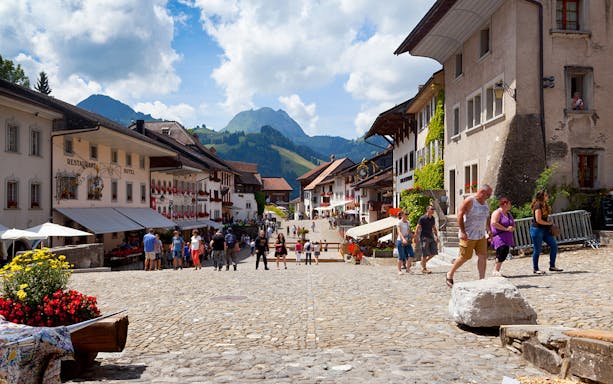 Cobbled street in Gruyères village with tourists, surrounded by traditional Swiss buildings and mountains.