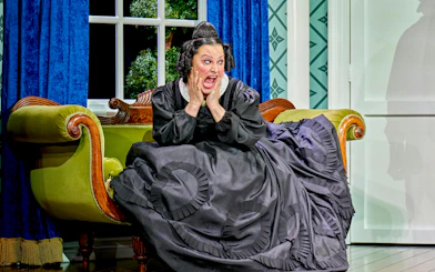 Woman in period costume on a vintage sofa, expressing surprise, from "Oh Mary!" show.