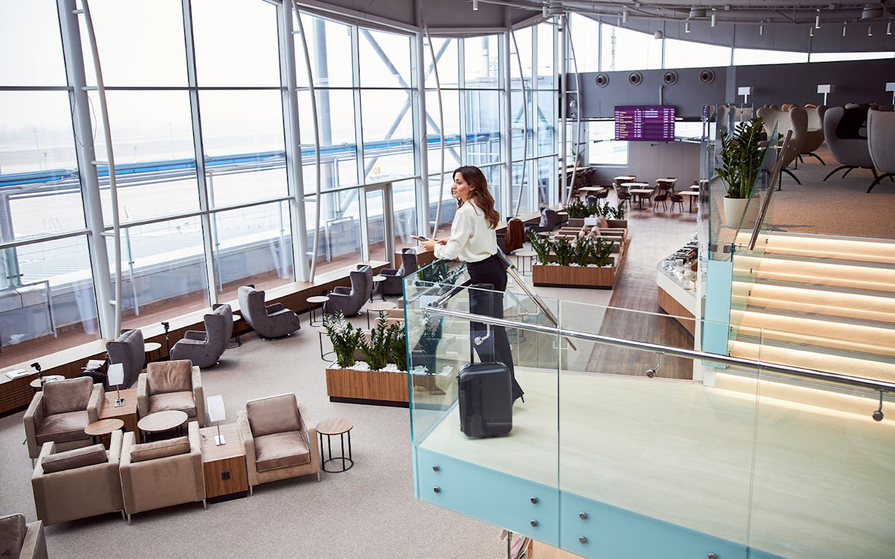 Woman standing in modern airport lounge with suitcase, overlooking seating area.
